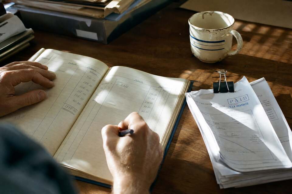 Hands entering figures into a hardcover ledger on a timber desk, beside a mug and a clipped stack of paid invoices, in afternoon light